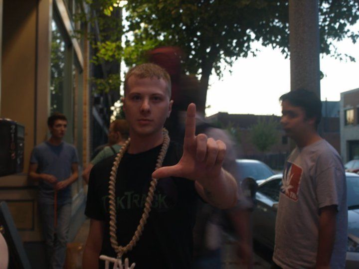 TakTrack in a thick rope chain making a hand sign at the camera on a city sidewalk.