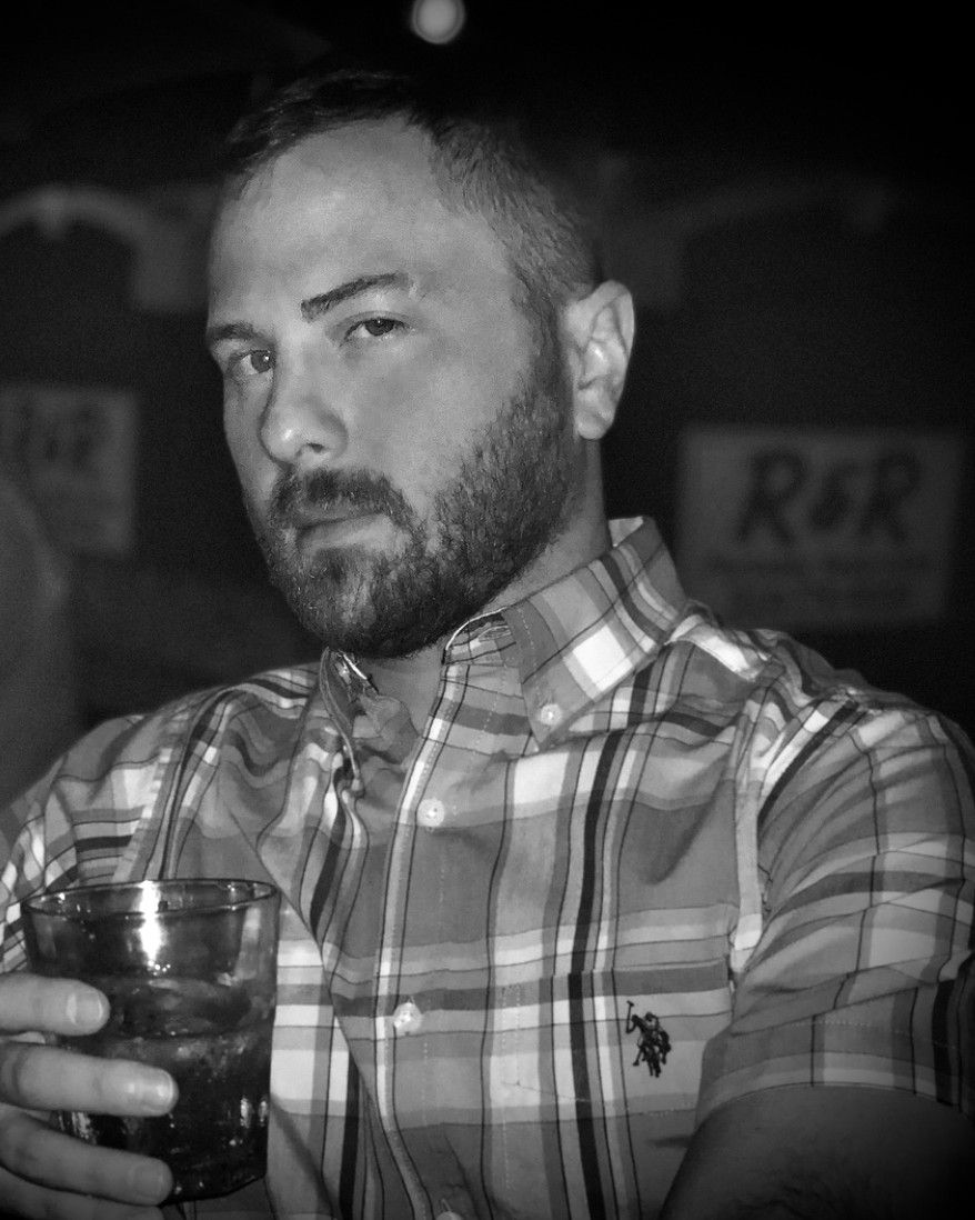 Black-and-white portrait of TakTrack with a beard, holding a glass.