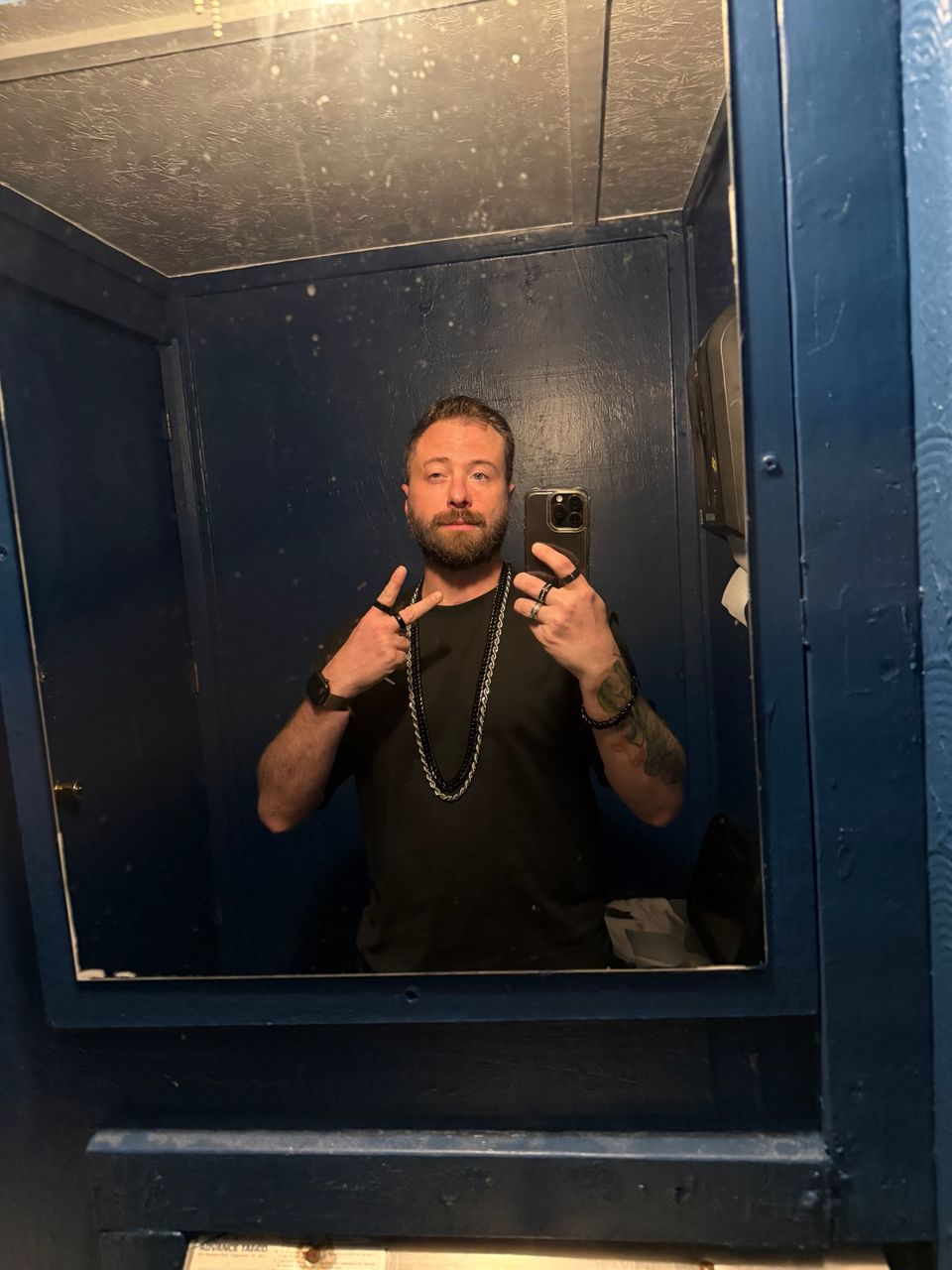 Mirror selfie of TakTrack in a black T-shirt, layered chains and rings, pointing at the camera in a small blue-painted room.
