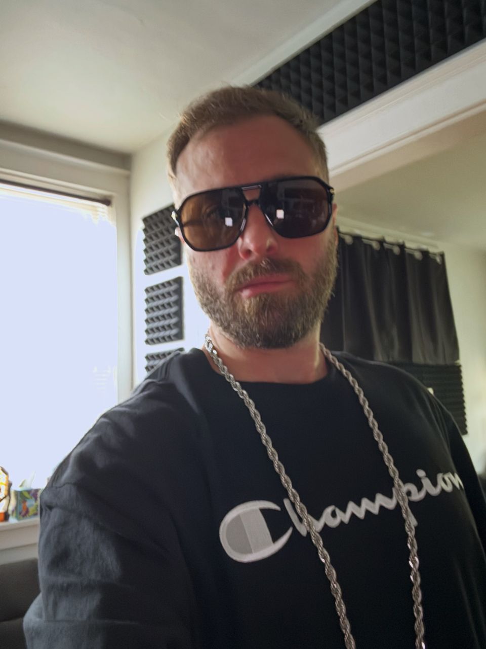 TakTrack in dark aviator sunglasses, a black Champion sweatshirt and a silver rope chain, in a room with foam acoustic panels on the walls.
