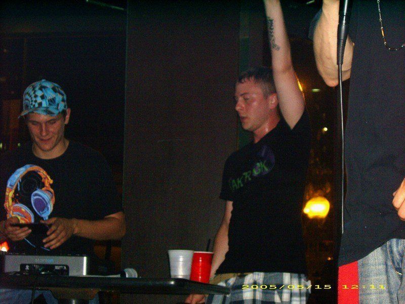 TakTrack raising an arm beside a DJ in a checkered cap at a DJ table in a dim club.