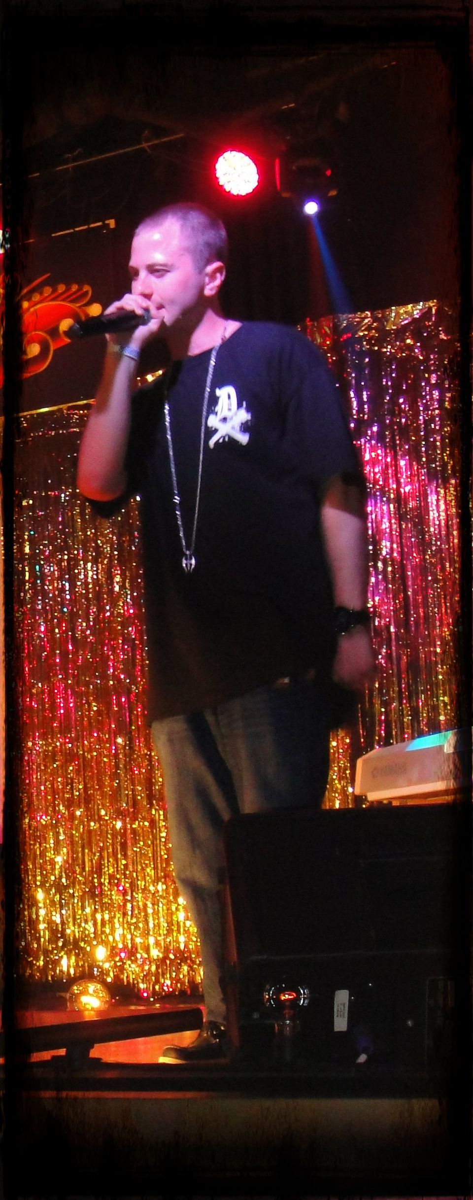 TakTrack performing on stage with a microphone in front of a sparkly gold curtain, wearing a black T-shirt.