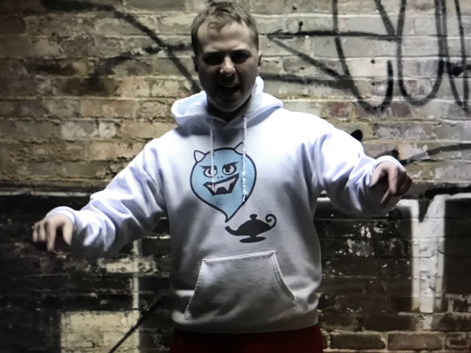 TakTrack in a white hoodie with a cartoon genie print, arms out against a graffiti-covered brick wall.