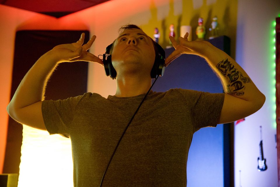 TakTrack in headphones and a gray T-shirt, hands raised beside his head, looking up in a red-lit studio.