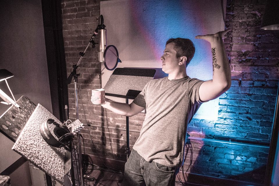 TakTrack in a gray T-shirt holding a mug, one arm raised, beside a microphone with a pop filter in a brick-walled vocal booth.