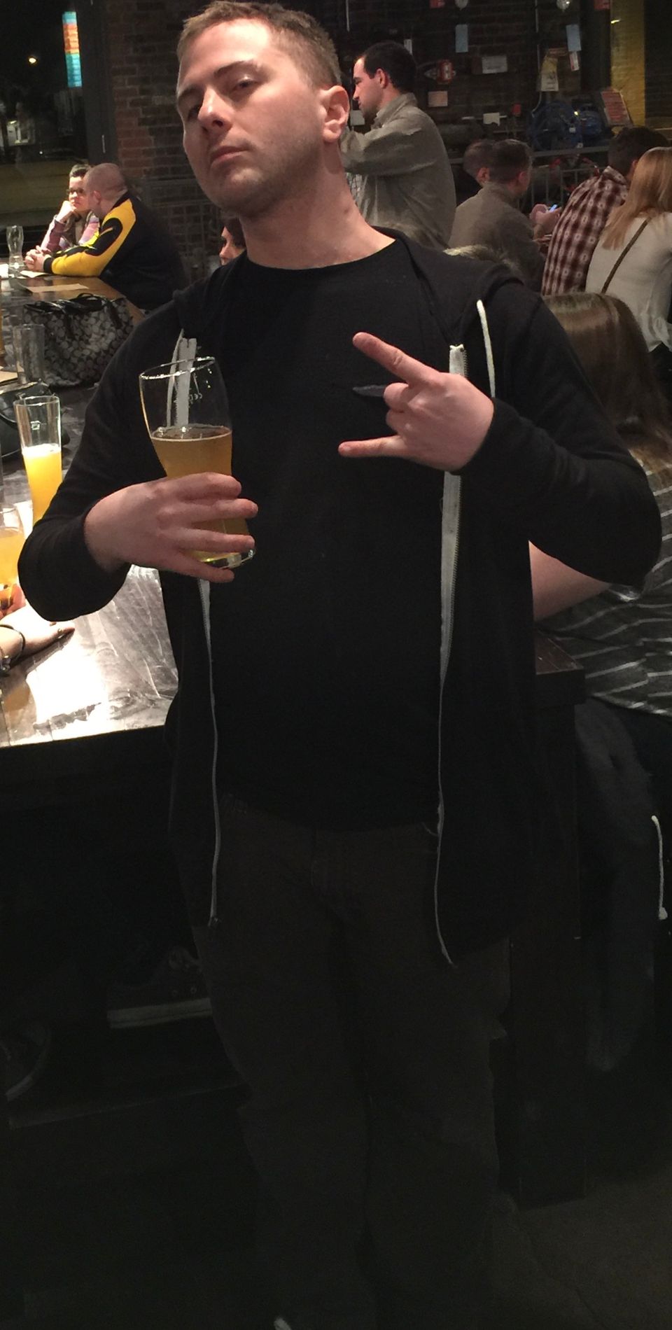 TakTrack holding a glass of beer and throwing up a hand sign in a crowded bar.