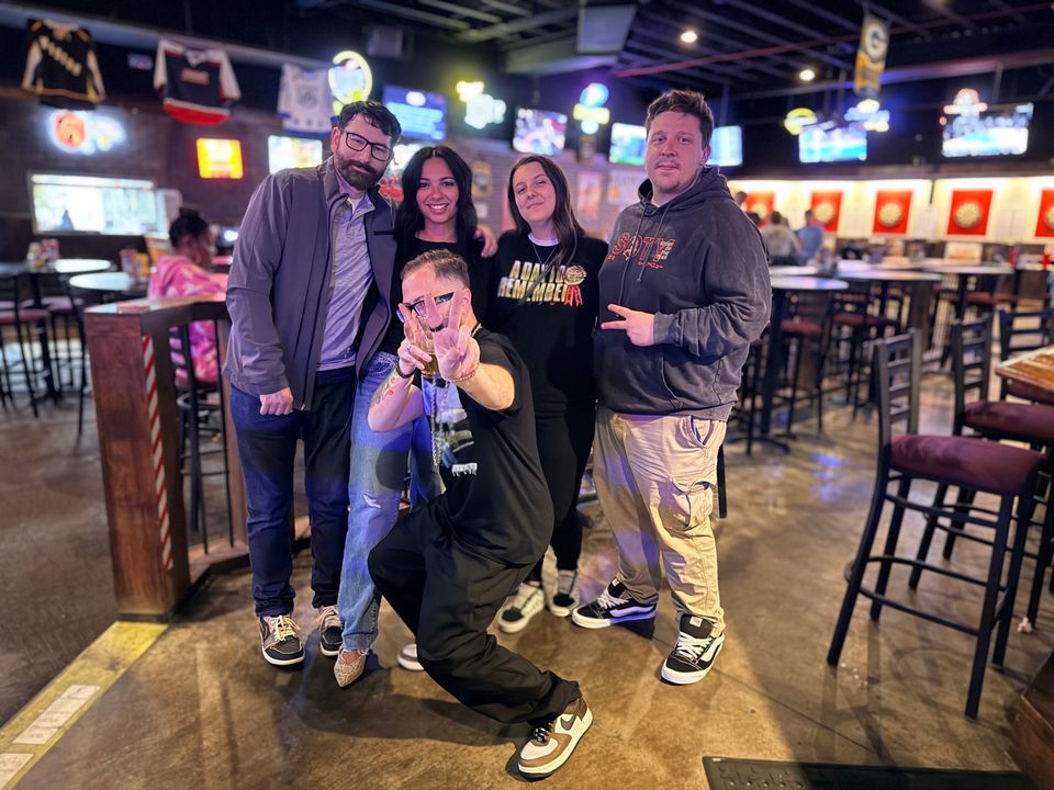 TakTrack kneeling in front of friends in a sports bar, pointing at the camera.