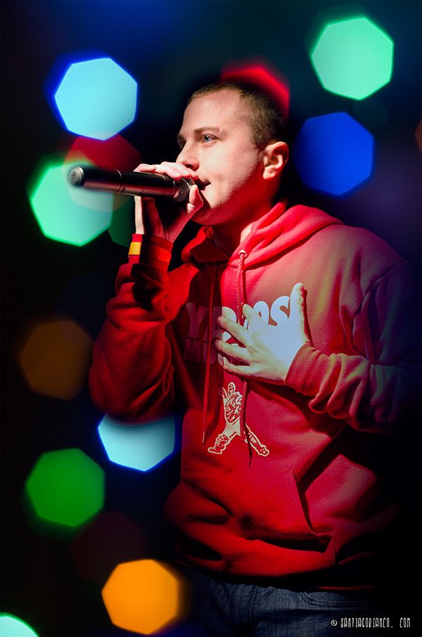TakTrack rapping into a microphone in a red hoodie, colored lights glowing behind him.