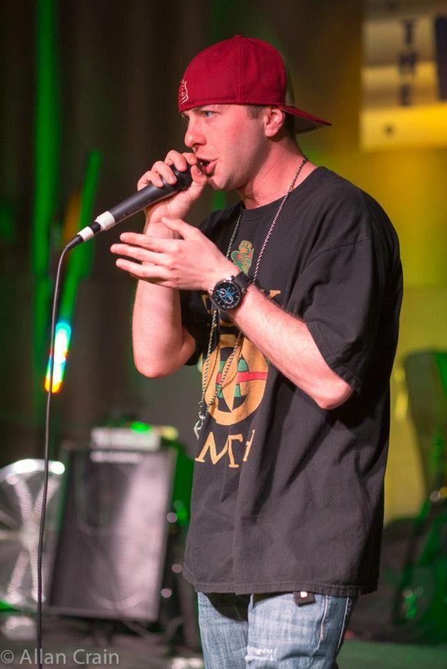 TakTrack performing on stage in a red cap and a black T-shirt, a microphone in one hand, a speaker behind him.