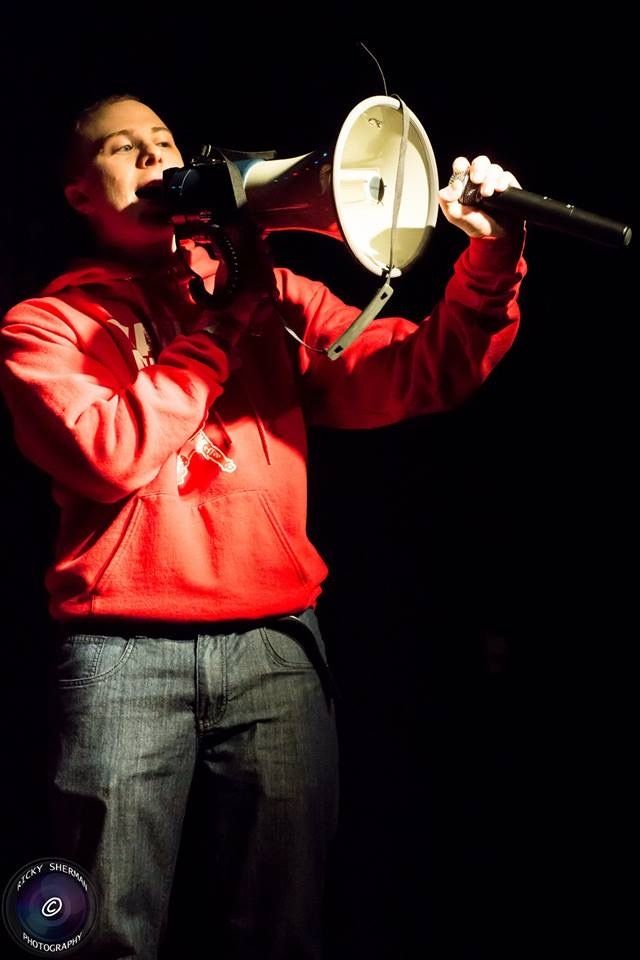 TakTrack in a red hoodie shouting through a megaphone on a dark stage, a microphone in his other hand.