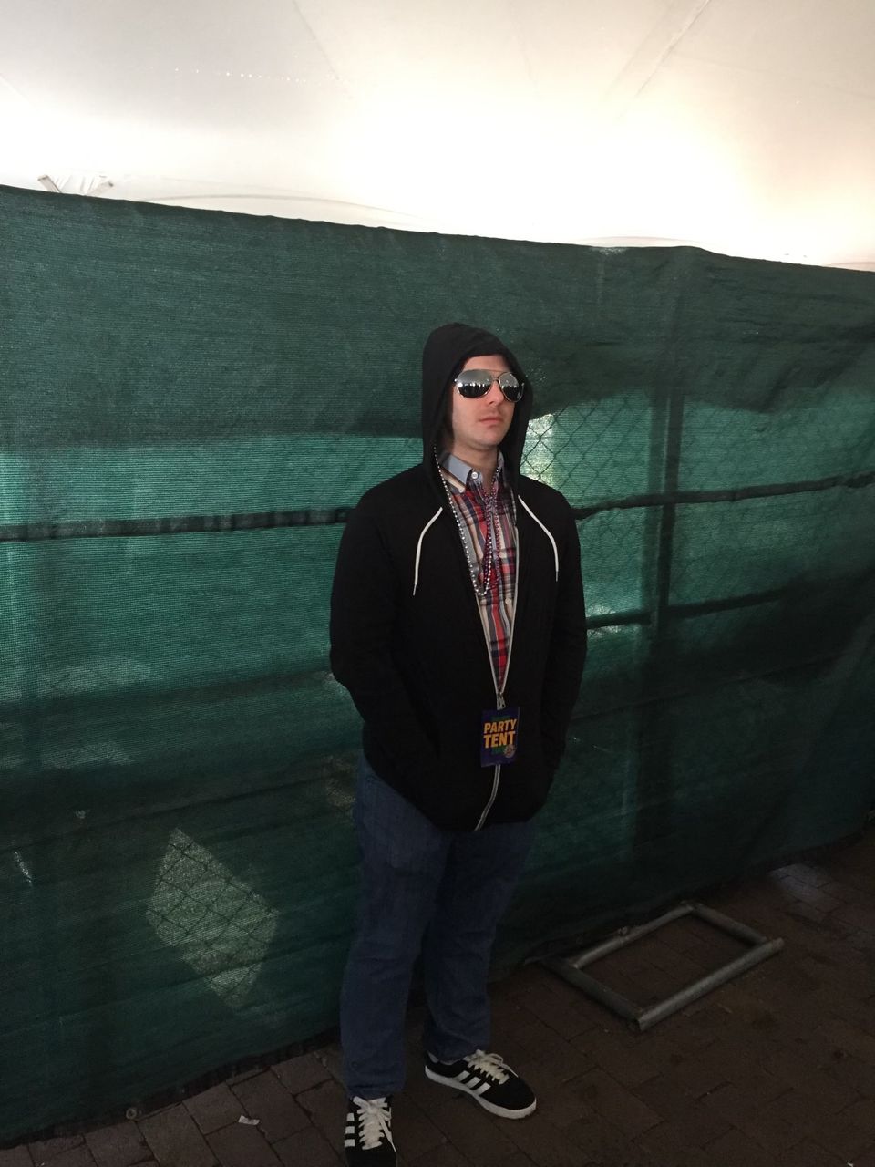 TakTrack in a black hoodie and sunglasses standing in front of a green fence.