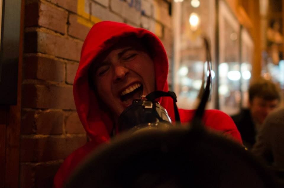 TakTrack in a red hoodie laughing in a brick-walled bar, a camera in the foreground.