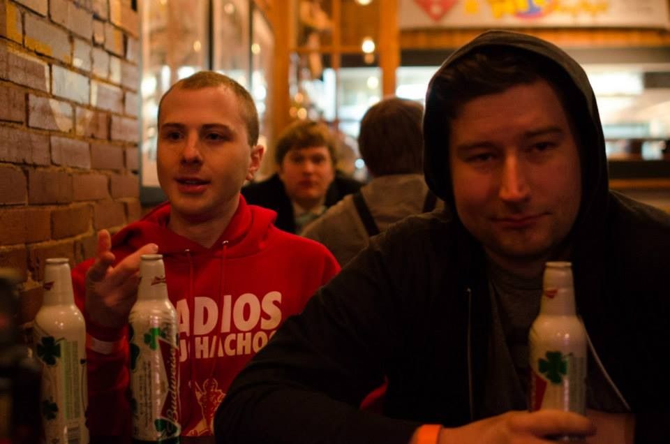 TakTrack in a red hoodie and a friend in a black hoodie at a bar, each holding a bottle.