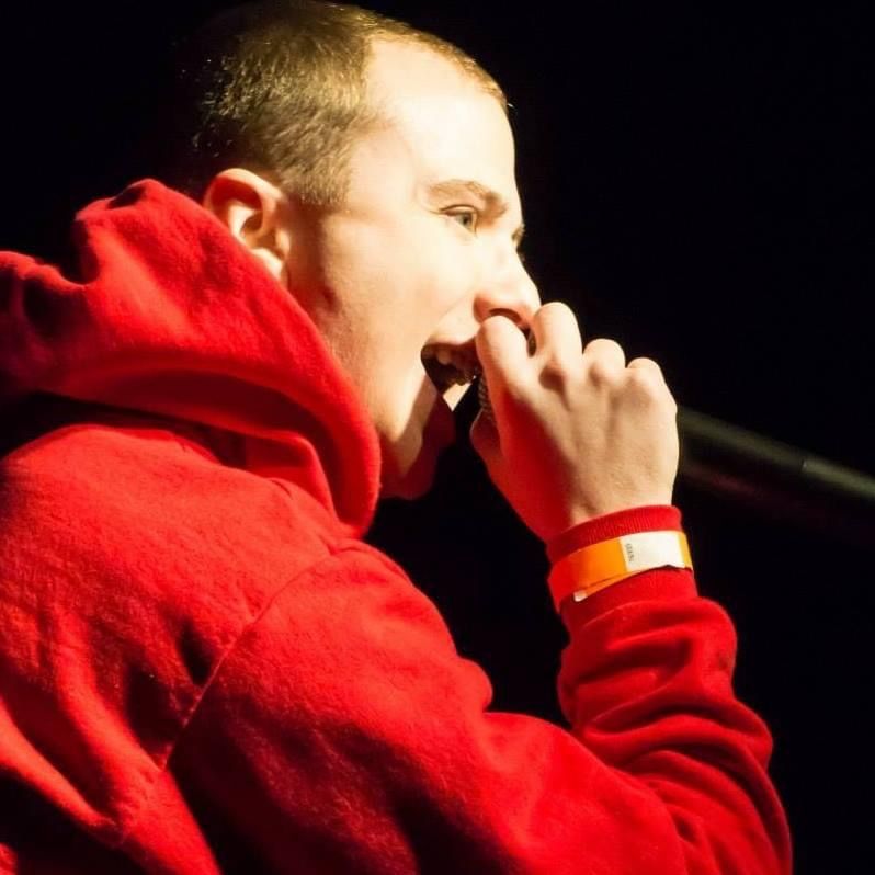Close-up profile of TakTrack in a red hoodie rapping into a microphone, an orange wristband on his wrist.