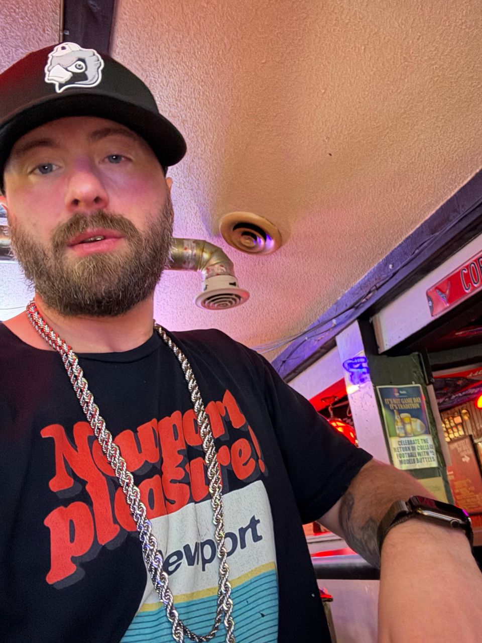 TakTrack in a black cap, a silver rope chain and a black graphic T-shirt, taking a selfie in a bar.